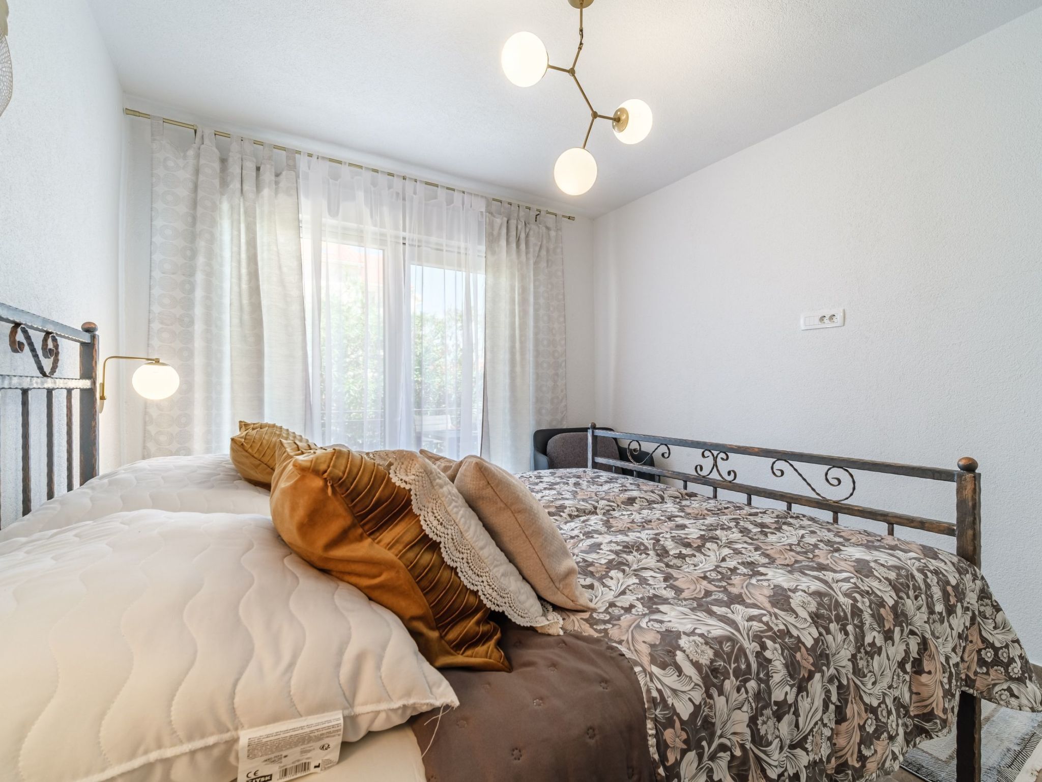 Un dormitorio con cama, una ventana con cortinas traslúcidas, una lámpara de pared y un enchufe.