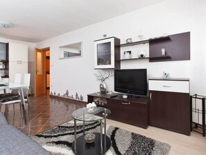 Ein modernes Wohnzimmer mit einem Glastisch, einem Flachbildfernseher, einer dunklen Holzkommode und einem Essbereich mit weißem Tisch und Stühlen.