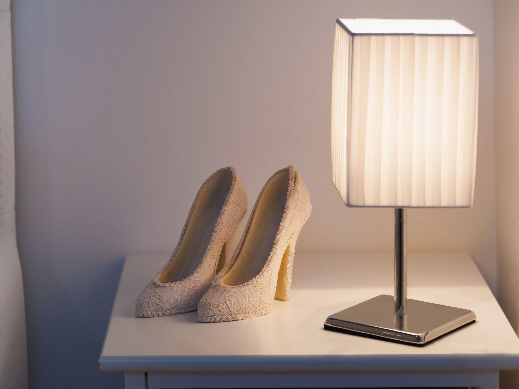 A pair of white lace high-heeled shoes on a white bedside table next to a modern lamp with a pleated shade.
