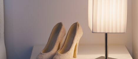 A pair of white lace high-heeled shoes on a white bedside table next to a modern lamp with a pleated shade.