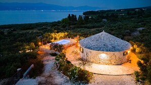 Una casa de piedra con techo circular, rodeada por un jardín con diversas plantas y flores, cerca de un cuerpo de agua bajo un cielo nocturno estrellado.