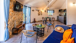 Amplia sala de estar con pared de piedra, televisor, mesa de comedor con sillas, zona de cocina y sofá azul.