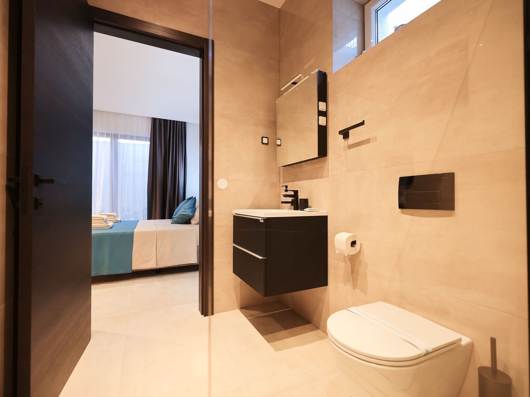 Un bagno moderno con servizi igienici, lavandino e specchio. Si intravede, attraverso una porta aperta sullo sfondo, un letto con copriletto blu e cuscini bianchi.