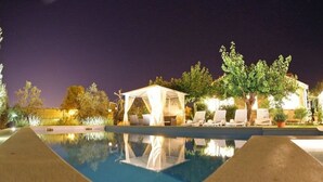 Ein beleuchteter Poolbereich mit überdachter Lounge und Liegestühlen.