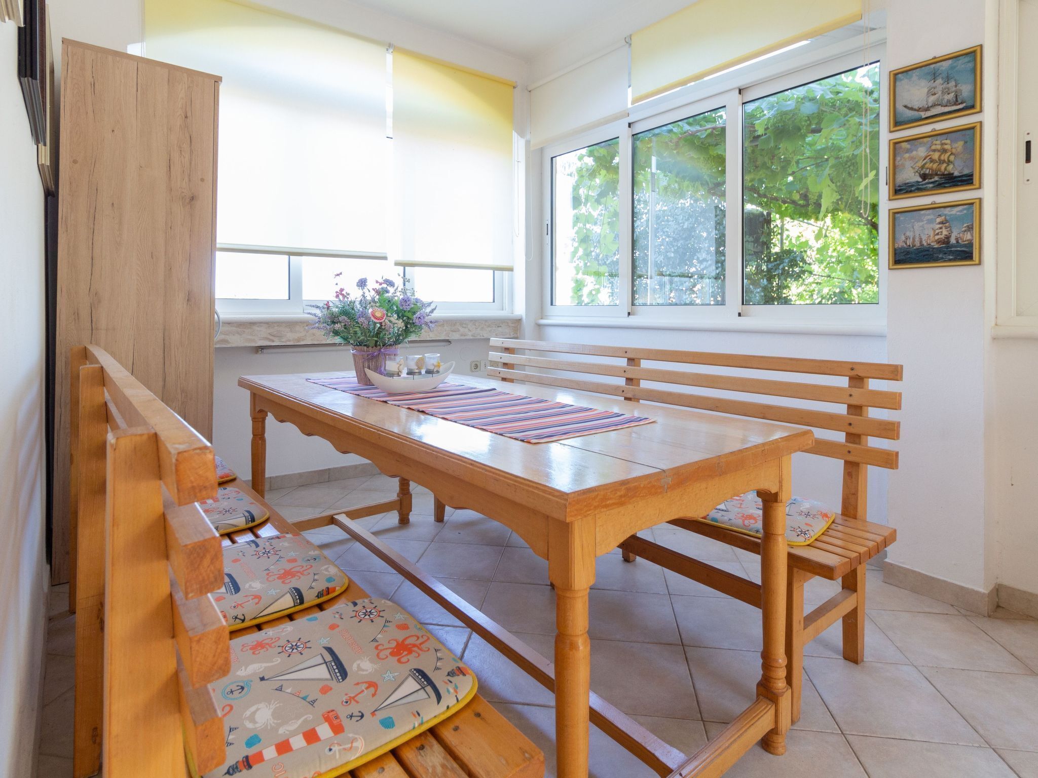 A dining area with a wooden table and benches, a window with a view of trees, and a wall with framed pictures.