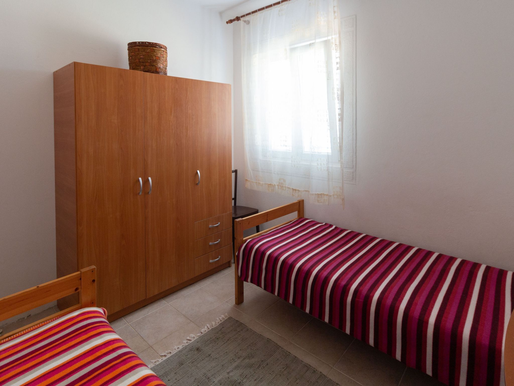 Un dormitorio con un armario de madera, una cama con colcha a rayas, una ventana con cortinas traslúcidas y una alfombra pequeña en el suelo.