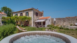 Una banyera d'hidromassatge al pati d'una casa amb mur de pedra, teulada d'àmples rajoles i jardí amb plantes i una pérgola.