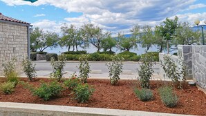 Un jardín ajardinado con diversas plantas y arbustos, un camino pavimentado y una vista a un cuerpo de agua y montañas al fondo.