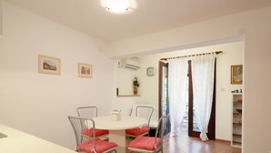 Un comedor con una mesa redonda y cuatro sillas, cada una con un cojín rojo. Hay dos cuadros en la pared, un reloj y una pequeña estantería con una planta.