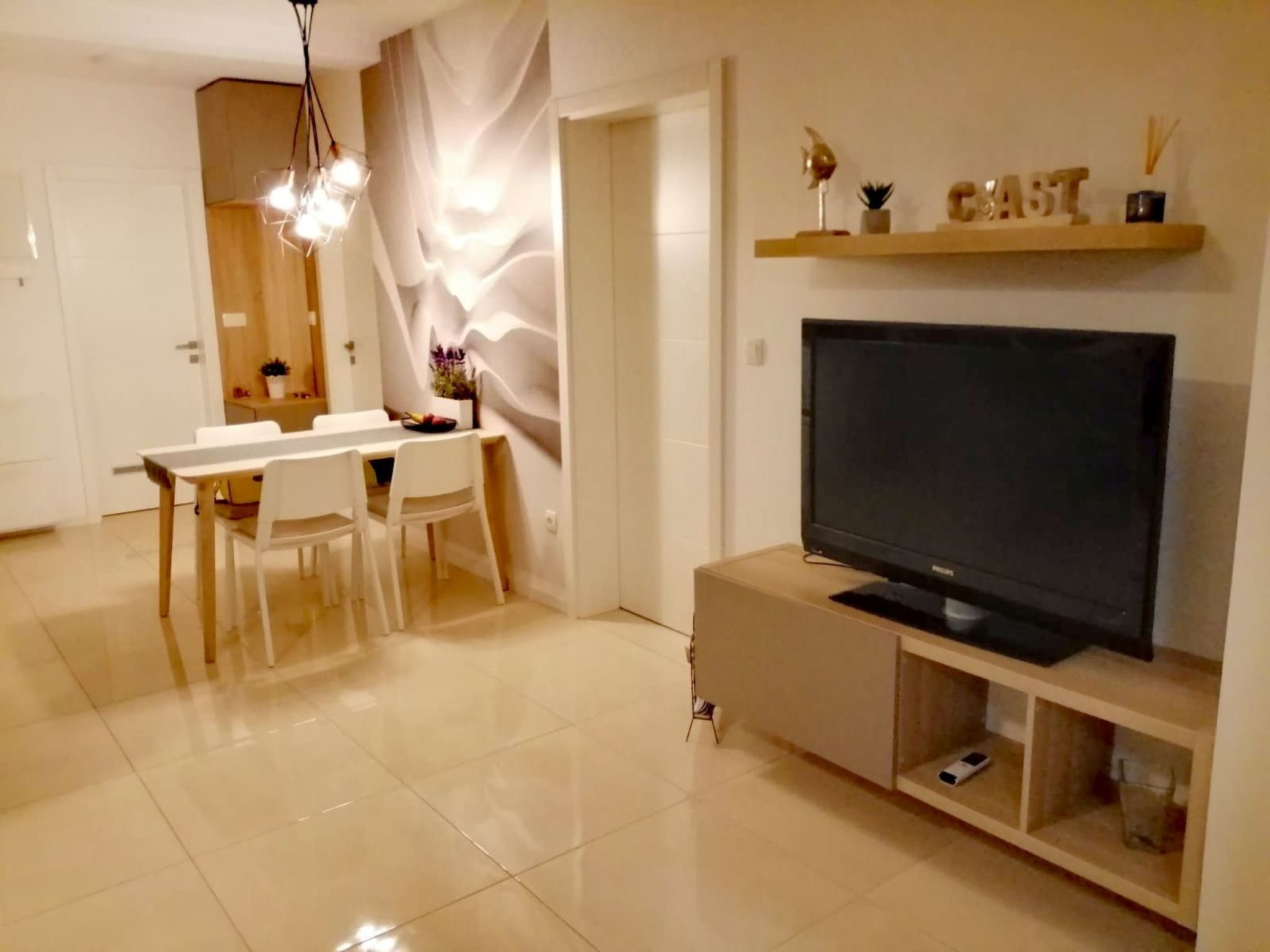A modern living room with a flat-screen TV, a dining area with a white table and chairs, and a decorative shelf with items.