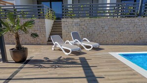 A poolside area with two white lounge chairs, a potted palm tree, and a stone wall with a staircase. - für 5 Personen ca. 68 m² in Šilo, Adriaküste Kroatien (Kroatische Inseln) by Interhome (Krk/Šilo)