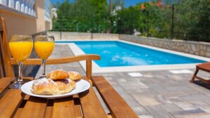A wooden table with a plate of pastries and two glasses of orange juice, overlooking a swimming pool. - Gemütliche Ferienwohnung Tina 4 by Interhome (Krk/Šilo)