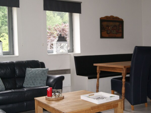 Een woonkamer met een zwarte leren bank, een houten salontafel, een houten eettafel met twee stoelen, en een raam met uitzicht op bomen.