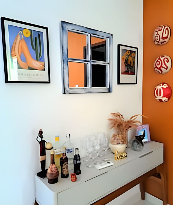 A room with a white desk, a window with a blue frame, and various decorative items including a vase, bottles, and framed artwork.