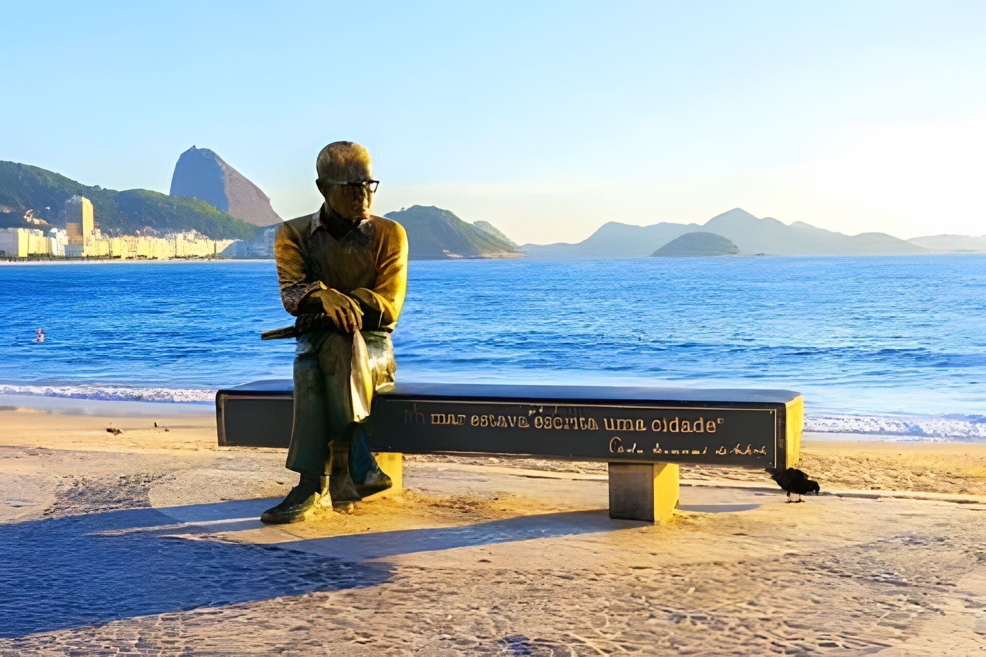 A statue of a man sitting on a bench with a plaque, overlooking a beach with the ocean and distant mountains.