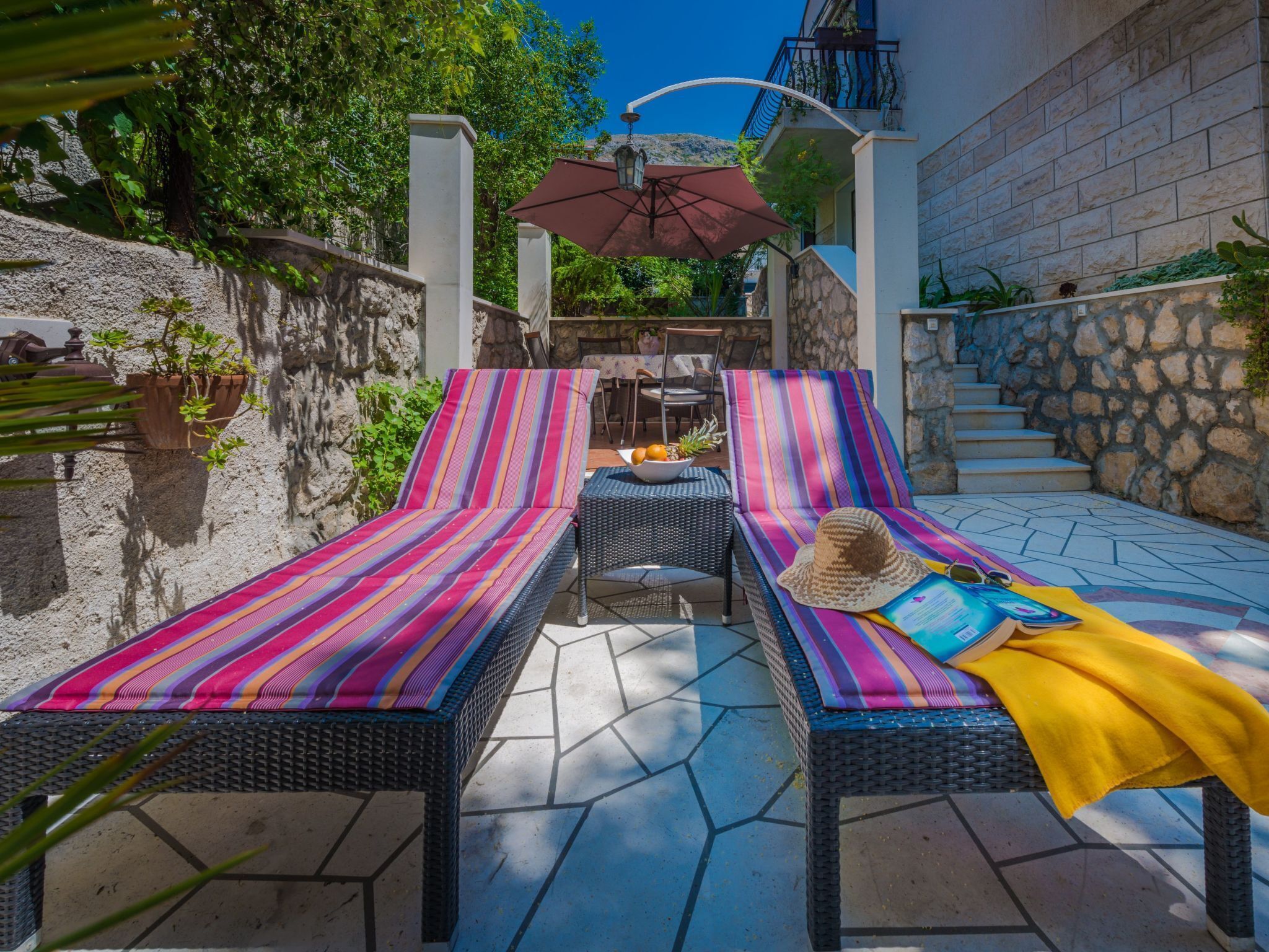 A sunlit outdoor seating area with two striped lounge chairs, a small table, and a decorative umbrella.