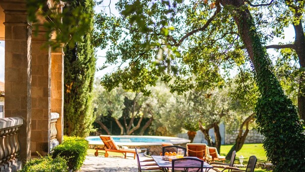 Un patio in pietra con tavolo e sedie, una piscina sullo sfondo e vegetazione lussureggiante.