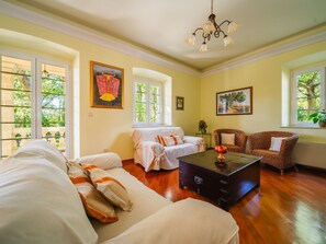 Ein geräumiges Wohnzimmer mit einer großen Couch, einem Couchtisch und zwei Korbstühlen. Zwei Fenster bieten Blick auf Grünflächen, und eine Chandelhier hängt von der Decke.