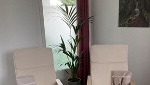 Duas poltronas bege com almofadas, uma pequena mesa de madeira com objetos decorativos, uma planta em vaso e um quadro na parede.