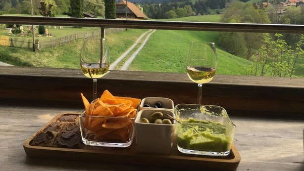 Une vue magnifique depuis une fenêtre donnant sur un paysage montagneux, avec une table en bois sur laquelle se trouvent un pain, des bâtonnets de carottes, des olives et une sauce verte, le tout accompagné de deux verres de vin blanc.