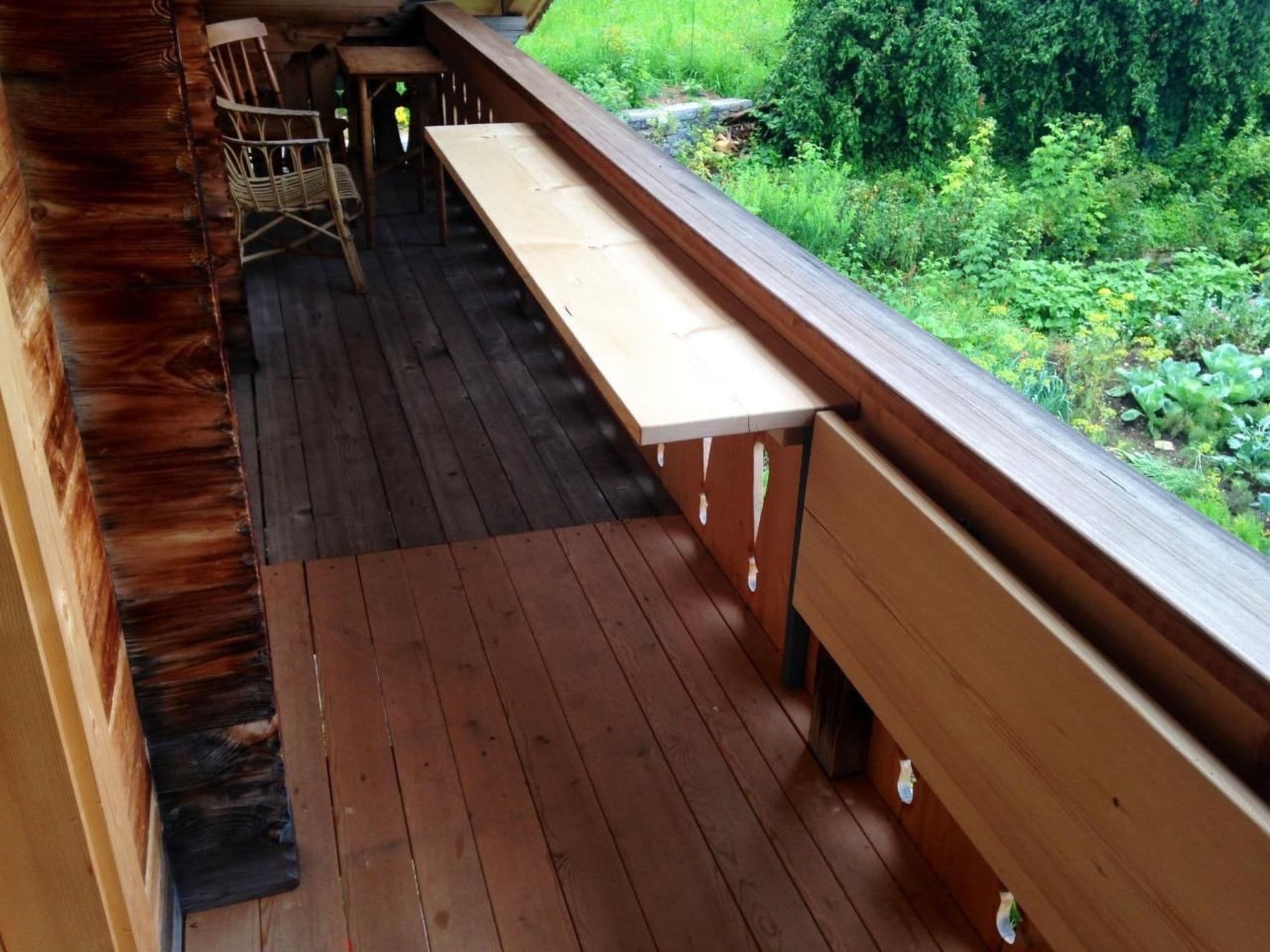 A wooden deck with a long bench and a table, surrounded by greenery.