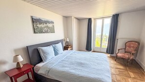 Une chambre à coucher avec un lit, une table de chevet, une chaise, une fenêtre avec des rideaux et un tableau au mur.