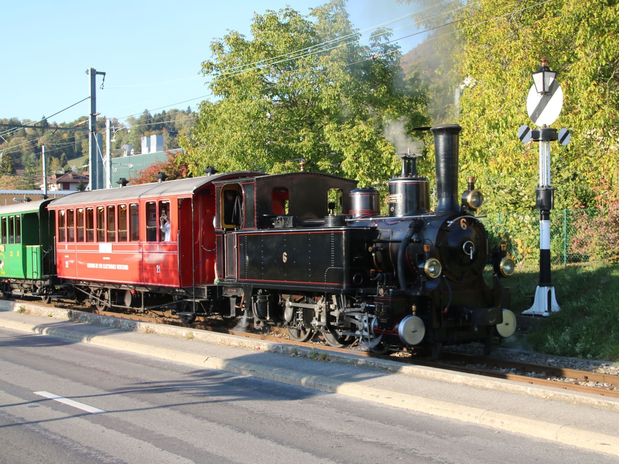 Une locomotive à vapeur d’époque, avec un wagon de voyageurs rouge et une locomotive noire, circule sur des voies ferrées situées à côté d’une route.