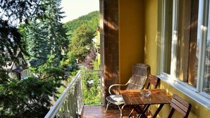 A balcony with a wooden table and chairs, a white railing, and a view of a green landscape with trees and a building in the background. - Daniela by Interhome (Carlsbad)