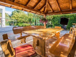 Ein hölzernes Pavillon im Freien mit Tisch und Bänken, umgeben von Grünpflanzen und einem Kiesweg.