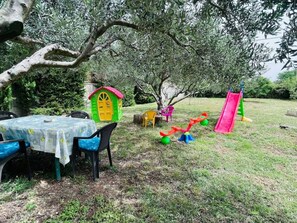 Ein grünes Areal im Freien mit einem Tisch, Stühlen, einem kleinen Spielhäuschen und einer Rutsche.