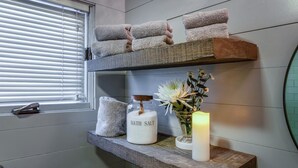 A bathroom with a window, a shelf with towels, a jar labeled 'BATH SALT,' a candle, and a vase with flowers. - Little Falls Bungalow (Brevard)
