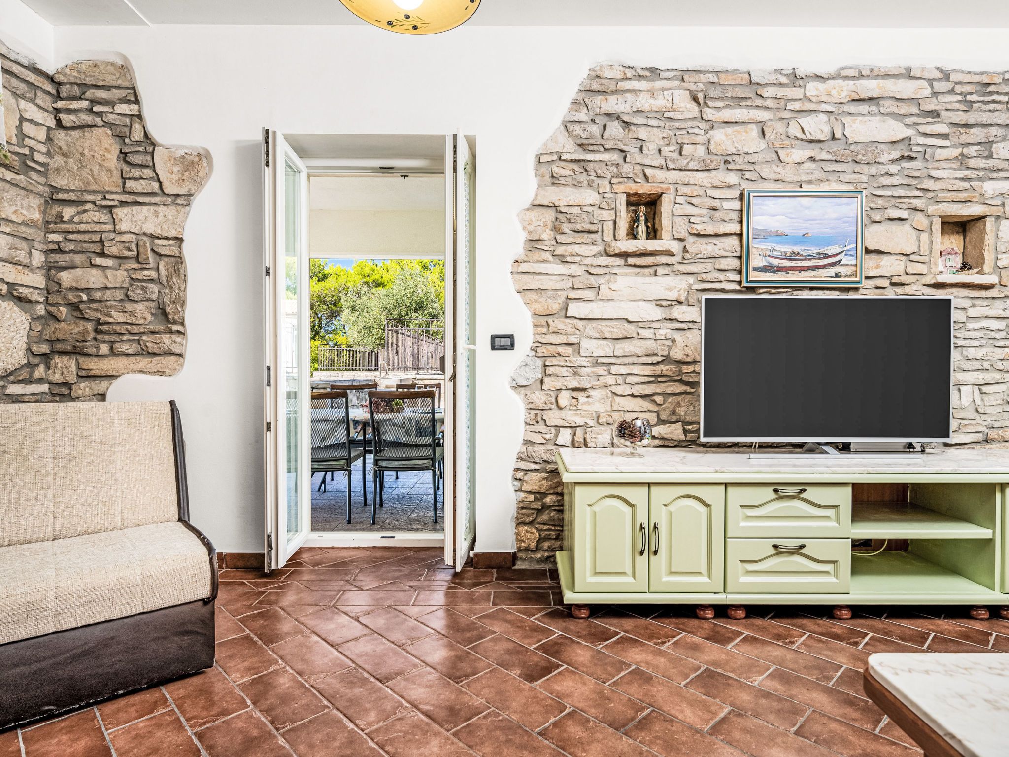 A room with a stone wall, a television on a cabinet, a sofa, and a door leading to an outdoor area with a table and chairs.