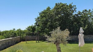 Un jardin avec un mur en pierre, une balançoire, un petit arbre et un parasol de patio.