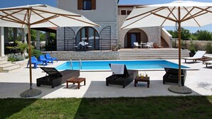 Un espace aménagé au bord de la piscine, avec des chaises longues, des parasols et un bâtiment en pierre en arrière-plan.