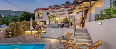 Una casa de dos pisos con piscina, mobiliario exterior y una escalera que lleva al piso superior.