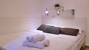 A neatly made bed with white linens, a brown pillow, and a patterned pillow. There are two wall-mounted lamps and a small potted plant on a shelf above the bed. - Studio Savic by Interhome (Krk/Šilo)