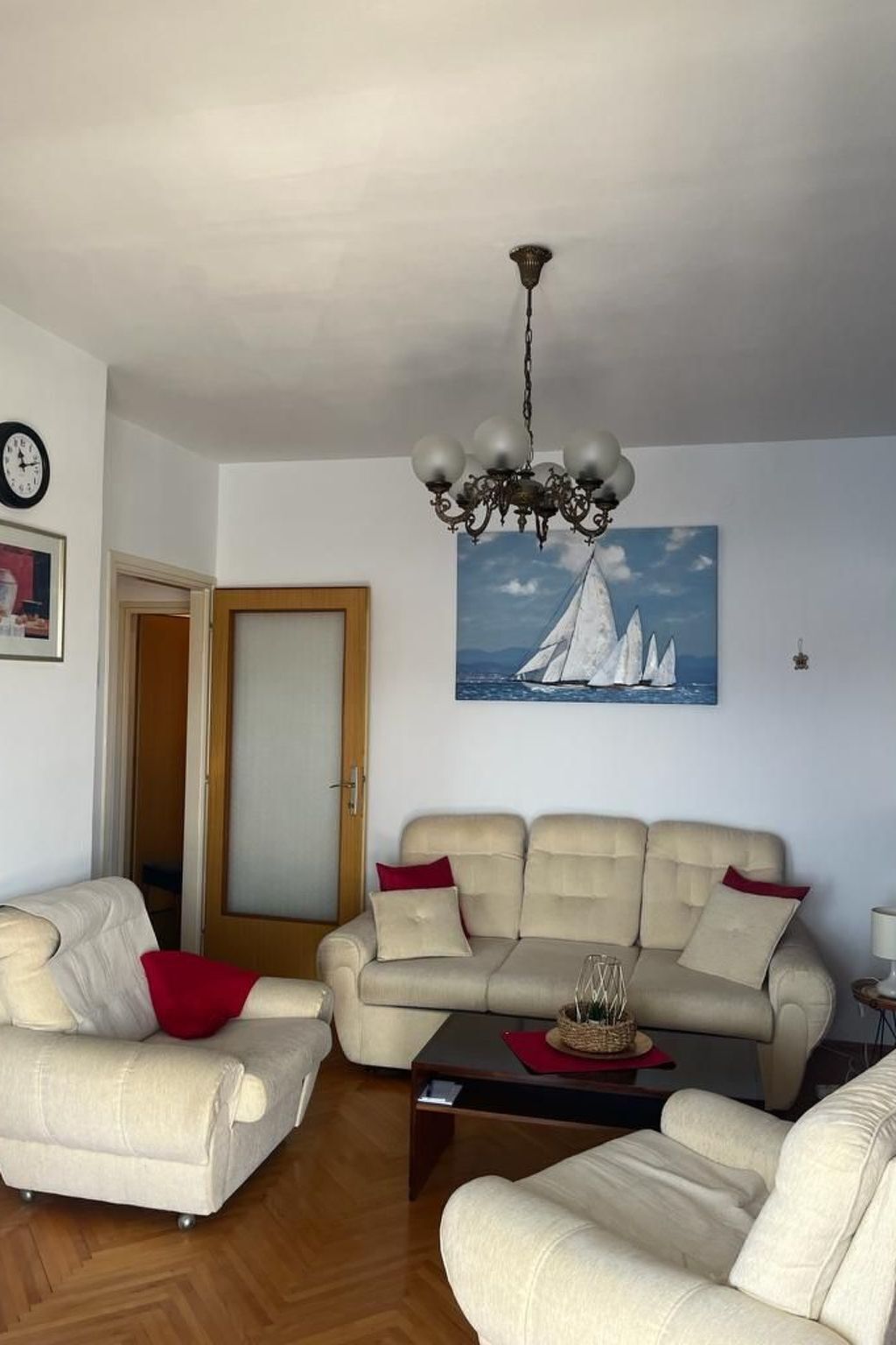 A living room with a beige sofa, a smaller armchair, a coffee table, and a chandelier. A painting of sailboats is on the wall.