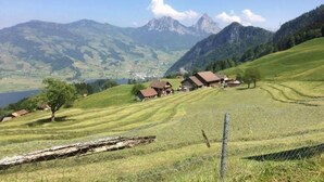 Paesaggio rurale con case su una collina, un corpo d'acqua e montagne sullo sfondo.