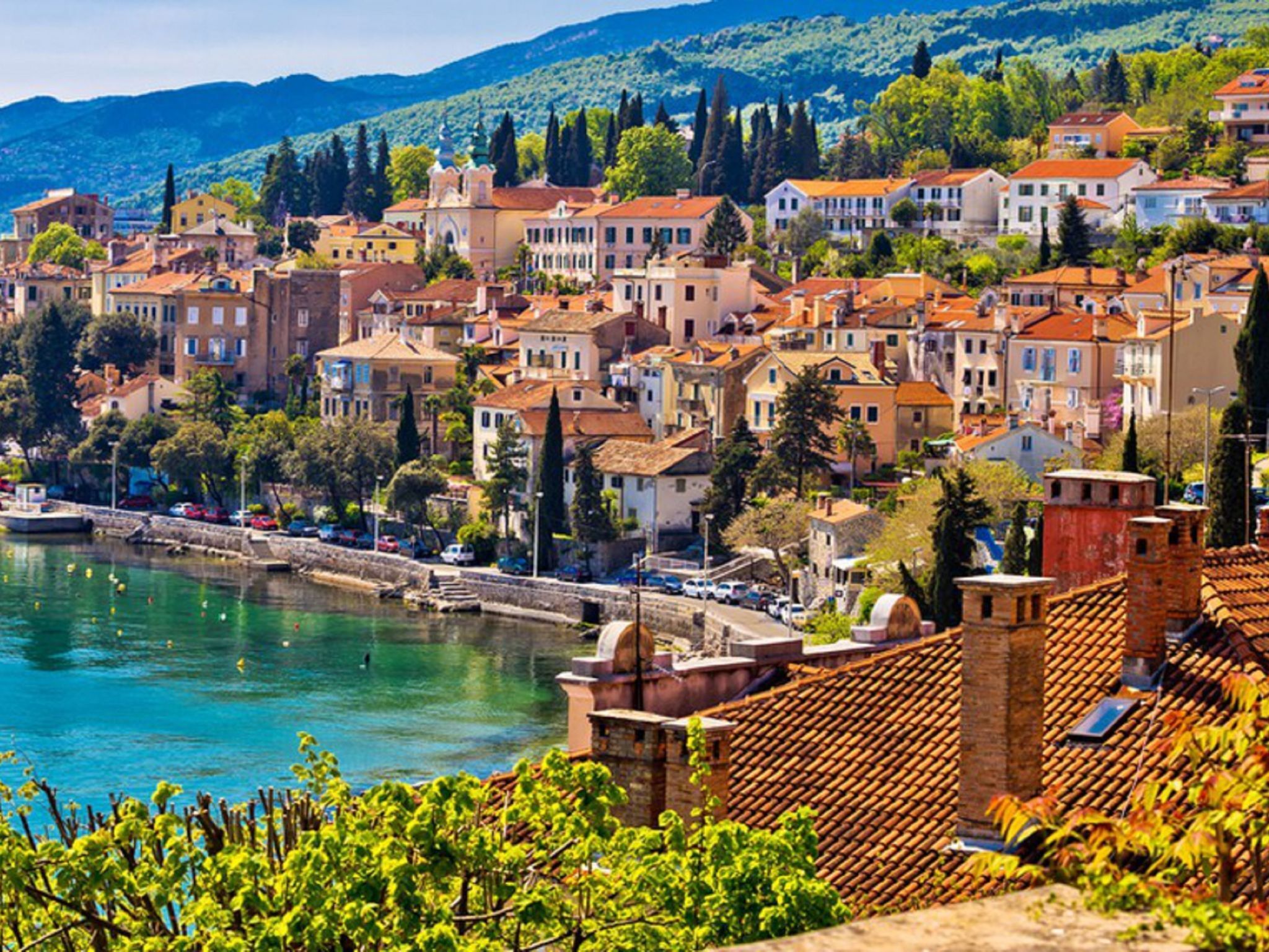 Une ville côtière aux bâtiments aux toits de tuiles rouges, dotée d’une promenade en bord de mer et d’une végétation luxuriante.