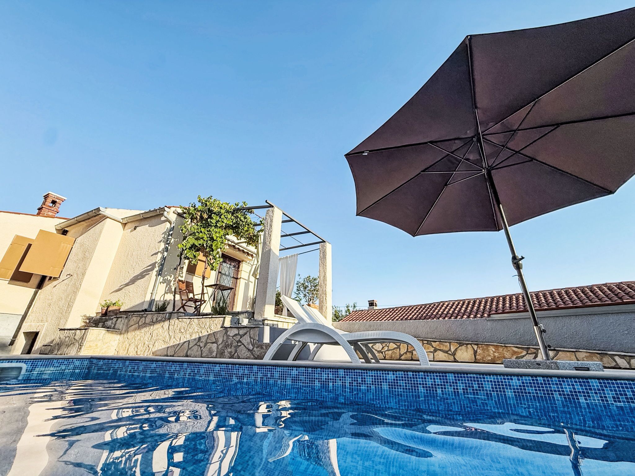 A swimming pool with a clear blue water surface, a large umbrella, and a building with a stone facade and a tiled roof.