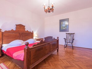 Ein hölzernes Bett mit roter Decke, ein Nachttisch aus Holz mit einer Lampe, ein Holzstuhl und ein Gemälde an der Wand.