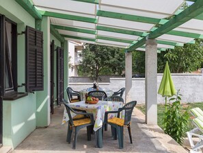Eine Terrasse im Freien mit Tisch und Stühlen, einem grünen Sonnenschirm und einer weißen Wand im Hintergrund.