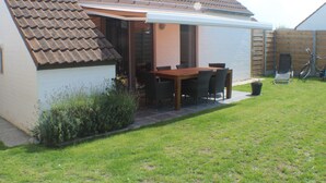 Ein Haus mit Ziegeldach, einer überdachten Terrasse mit Tisch und Stühlen, einem Blumentopf und einem Fahrrad im Garten.