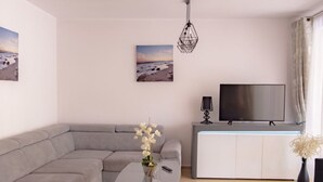 En moderne stue med en grå hjørnesofa, et glas sofabord, et fladskærms TV på et hvidt skab og en hængende lampe.