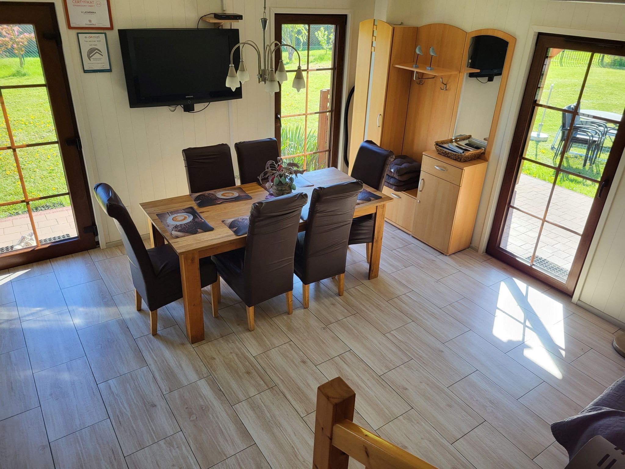 Un comedor con una mesa y sillas de madera, un televisor de pantalla plana montado en la pared y vistas al jardín a través de puertas de vidrio.