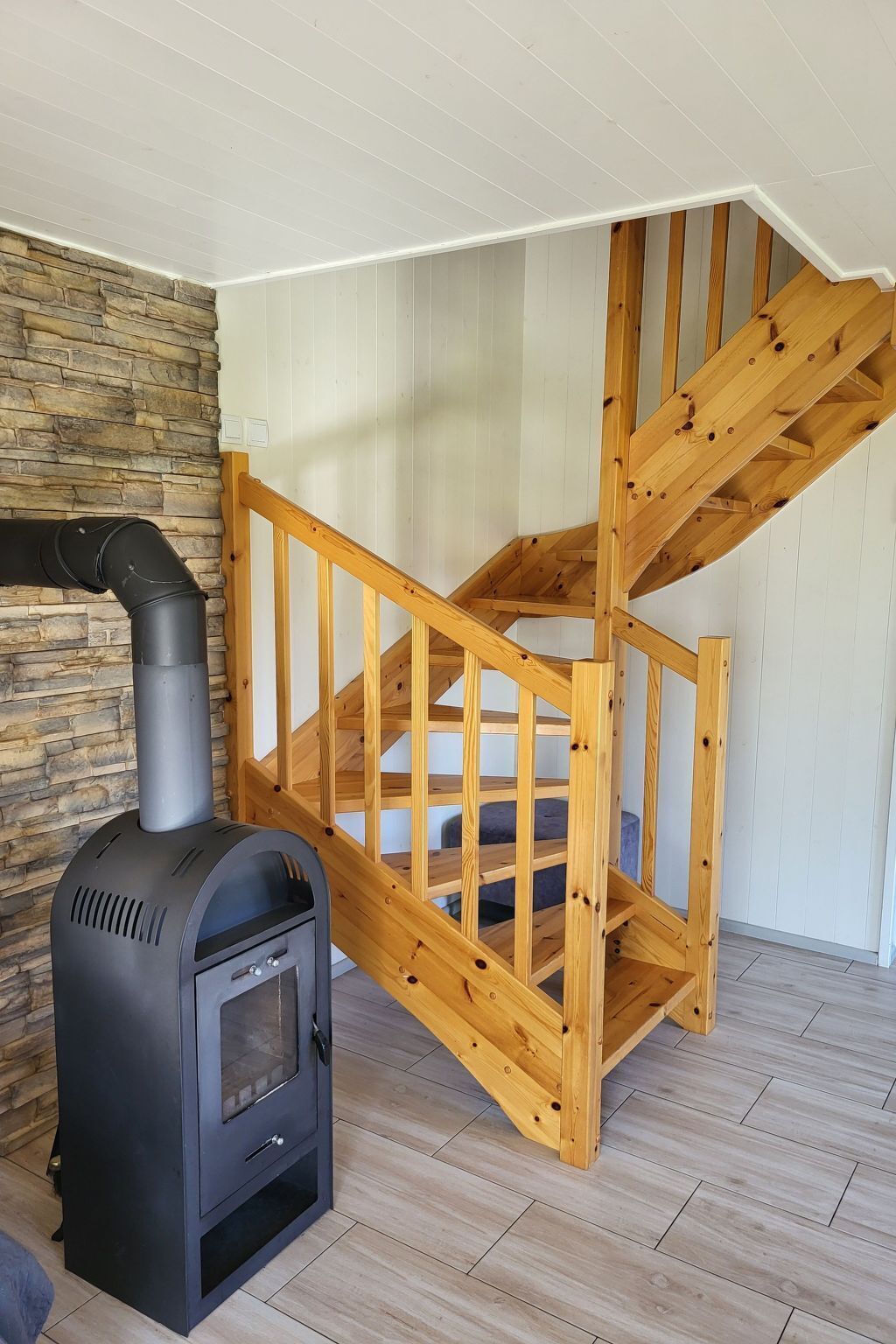 A wooden staircase with a black wood-burning stove in a room with white walls and a stone accent wall.