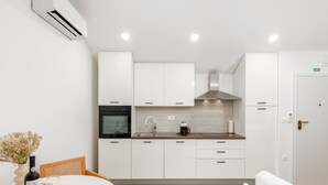 Una cocina moderna con armarios blancos, horno negro, campana extractora de acero inoxidable y un lavamanos. Hay un comedor con una mesa y sillas, y un sofá blanco.