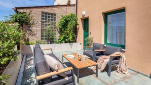 Un patio exterior con un sofá, sillas y una mesa. Se observa una pared de ladrillo con una ventana y follaje.