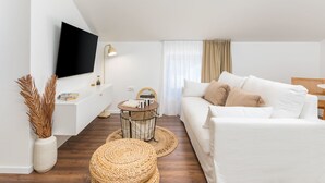 Una sala de estar moderna con un sofá blanco, un televisor de pantalla plana montado en la pared, una mesita redonda con una lámpara y un pouf tejido.