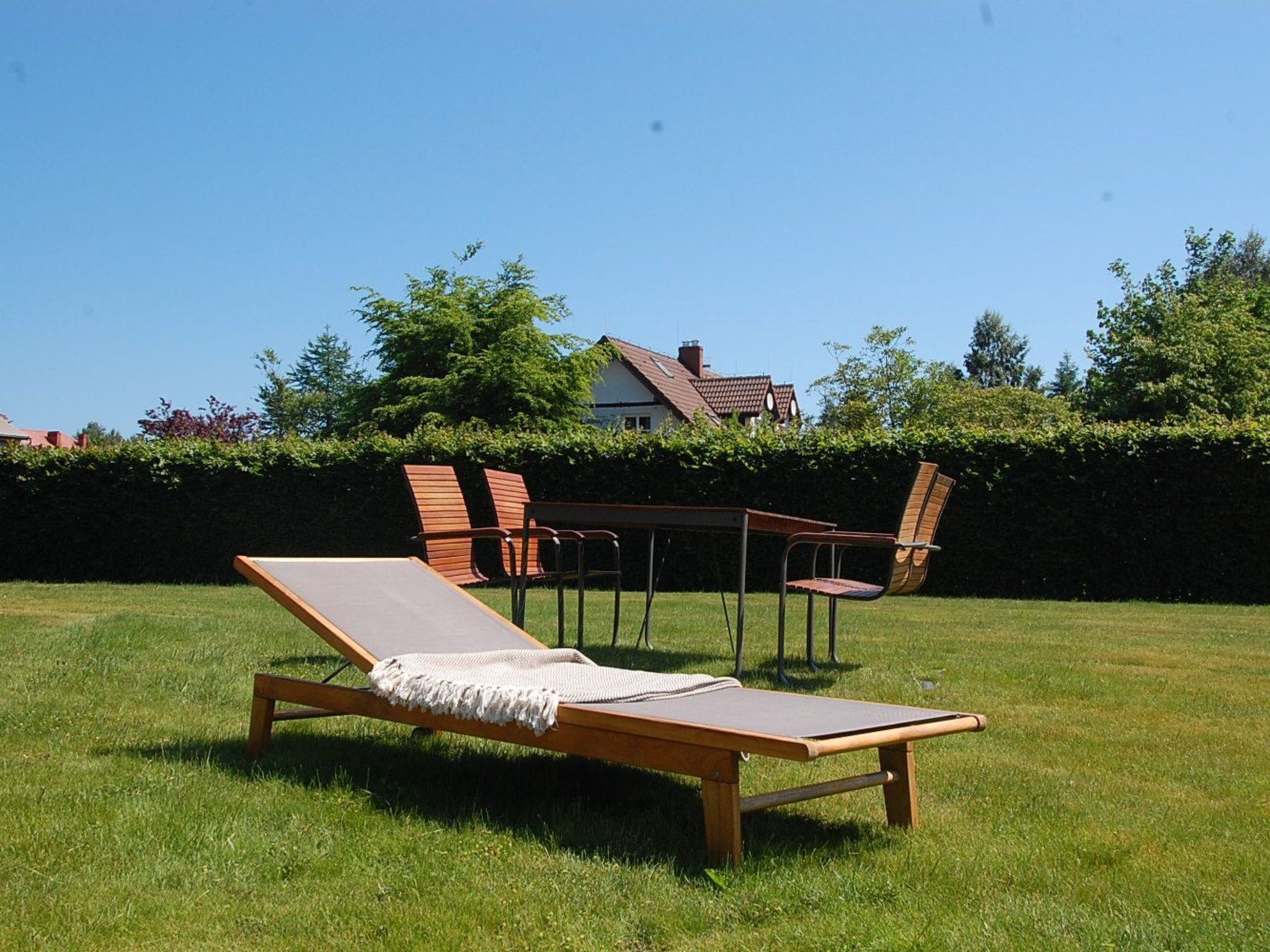 Un fauteuil de jardin en bois avec un coussin, ainsi qu’une table entourée de chaises, le tout installé dans un espace extérieur verdoyant.